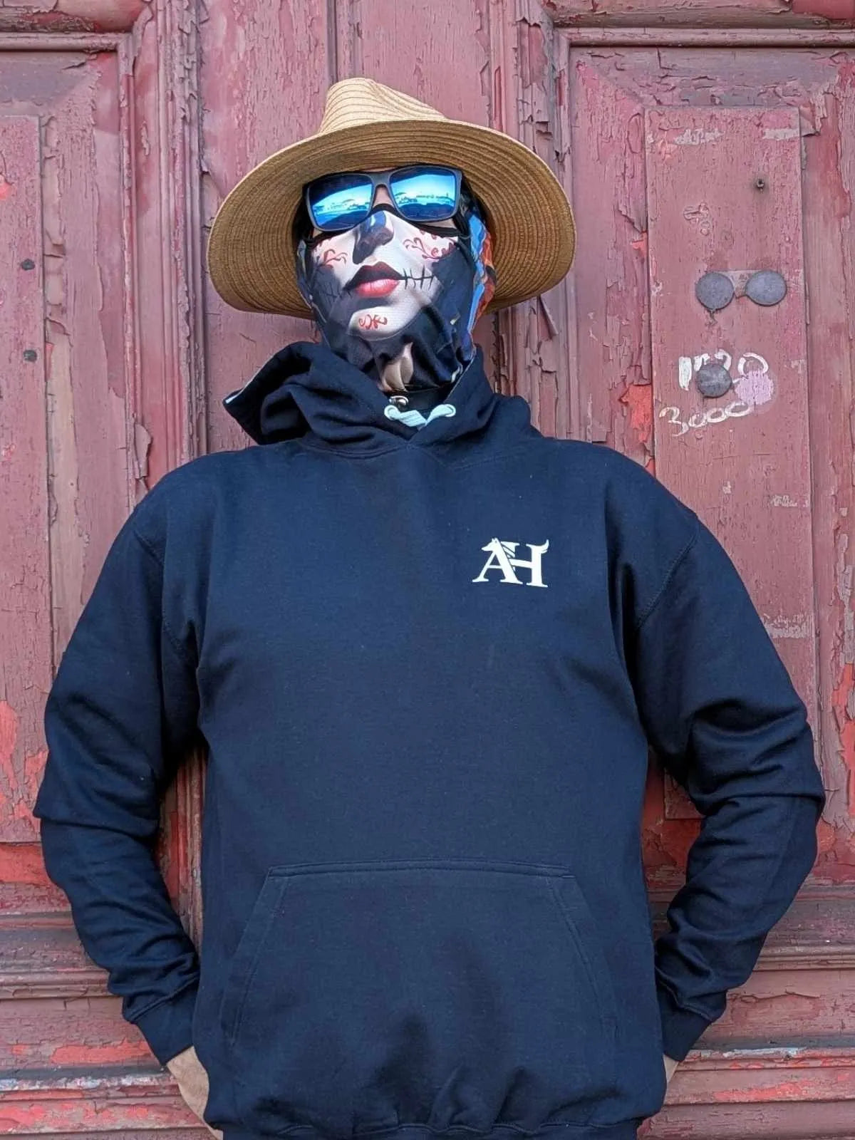 The Sun Walker wearing an Albert Harry AH minimal Dire Wolf Clan navy hoodie, sunglasses, and a straw hat against the red wooden door.