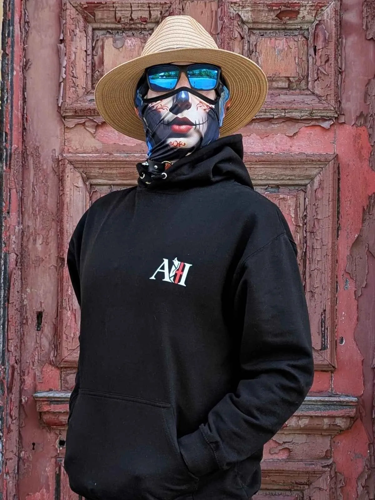 The Sun Walker, wearing an Albert Harry AH minimal White Knight black hoodie, sunglasses, and a straw hat, stood against the red wooden door.