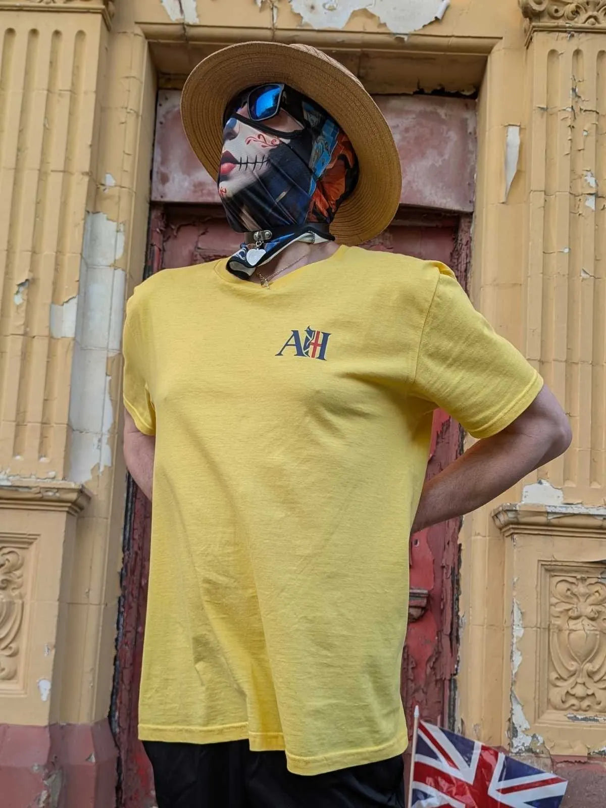 The Sun Walker, wearing an Albert Harry AH minimal White Knight yellow haze t-shirt, sunglasses, and a straw hat, stood against the red wooden door of the Trafoord Park Hotel.