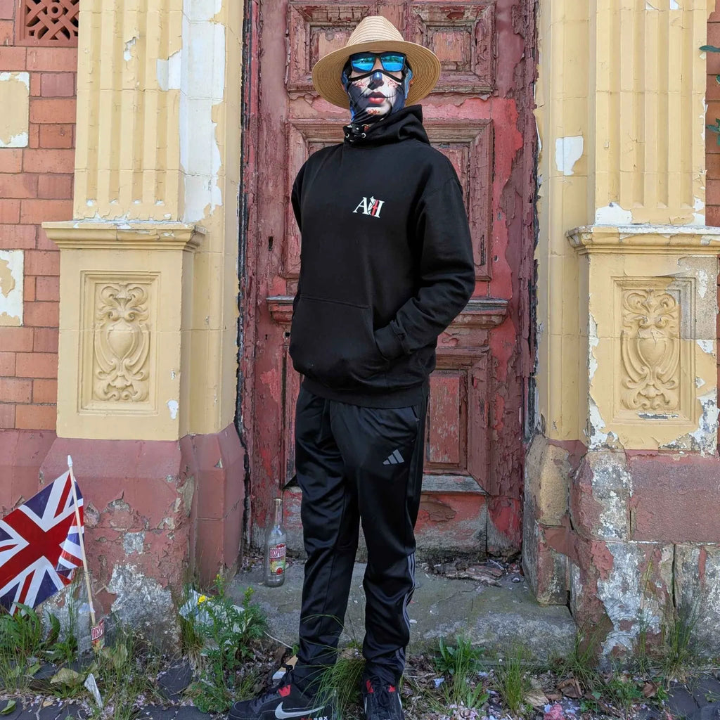 The Sun Walker, wearing an Albert Harry AH minimal White Knight black hoodie, sunglasses, and a Summer straw hat, stood against the red wooden door, Trafoord Park Hotel entrance.